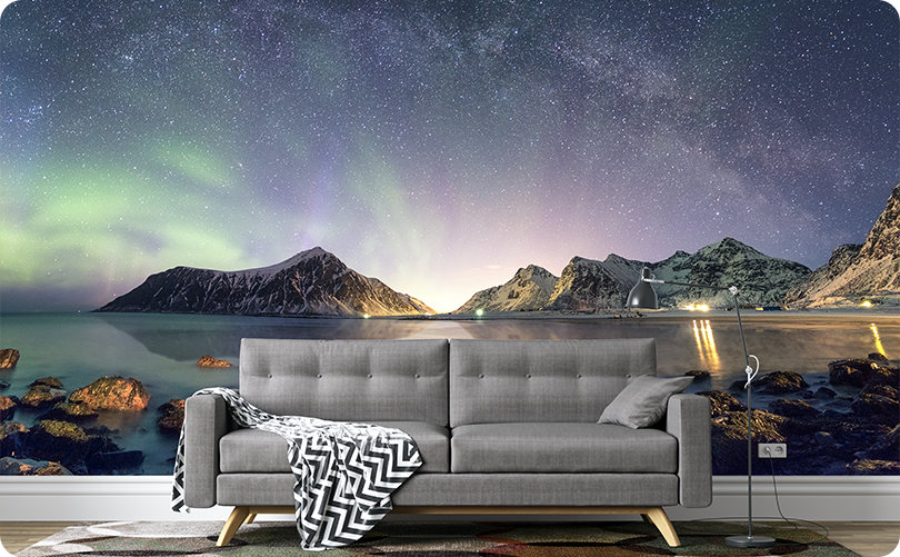 Green aurora and starry sky wallpaper mural with mountains and a rocky shoreline behind a gray sofa